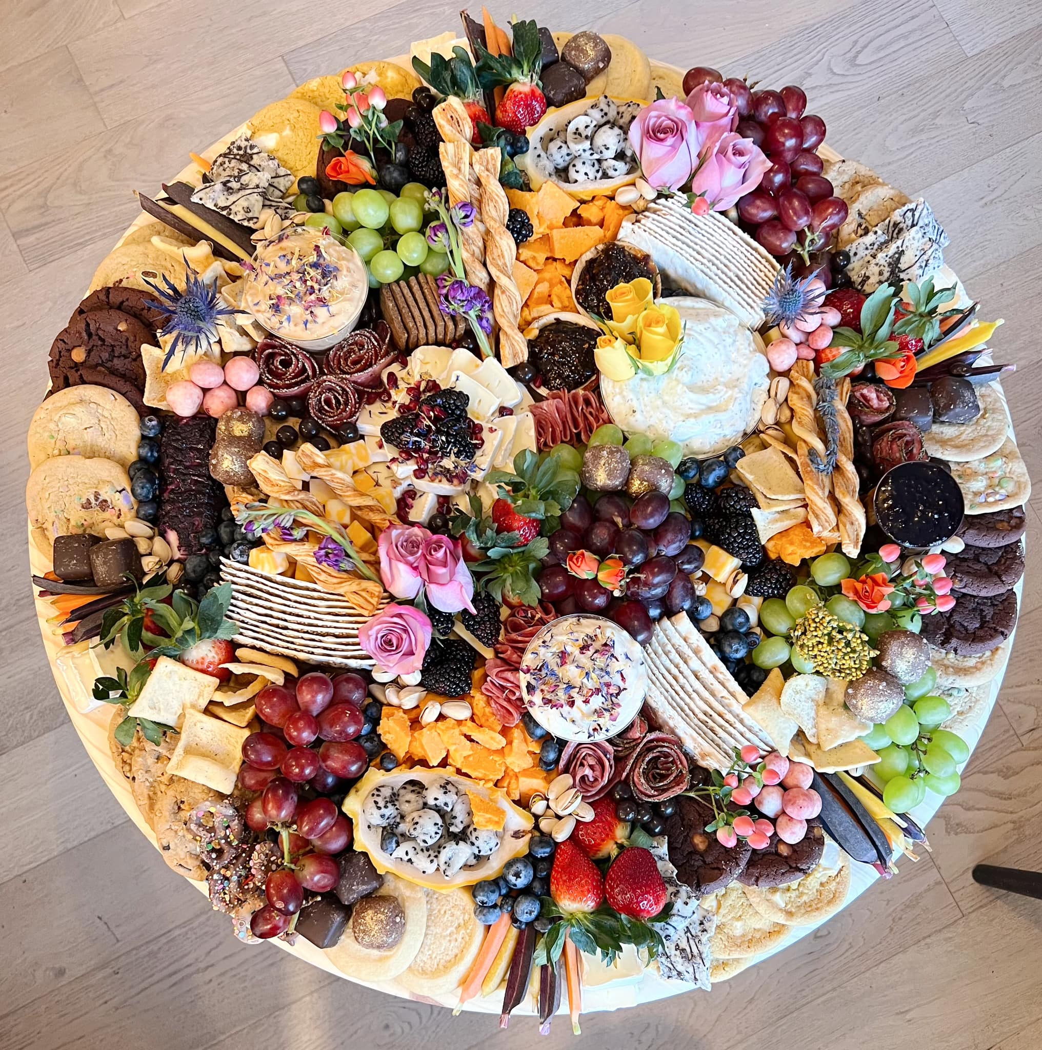 An overhead view of a round grazing board — pink roses, dragonfruit, grapes, dipped pretzels, cheeses, and chocolate cookies arranged in abundance