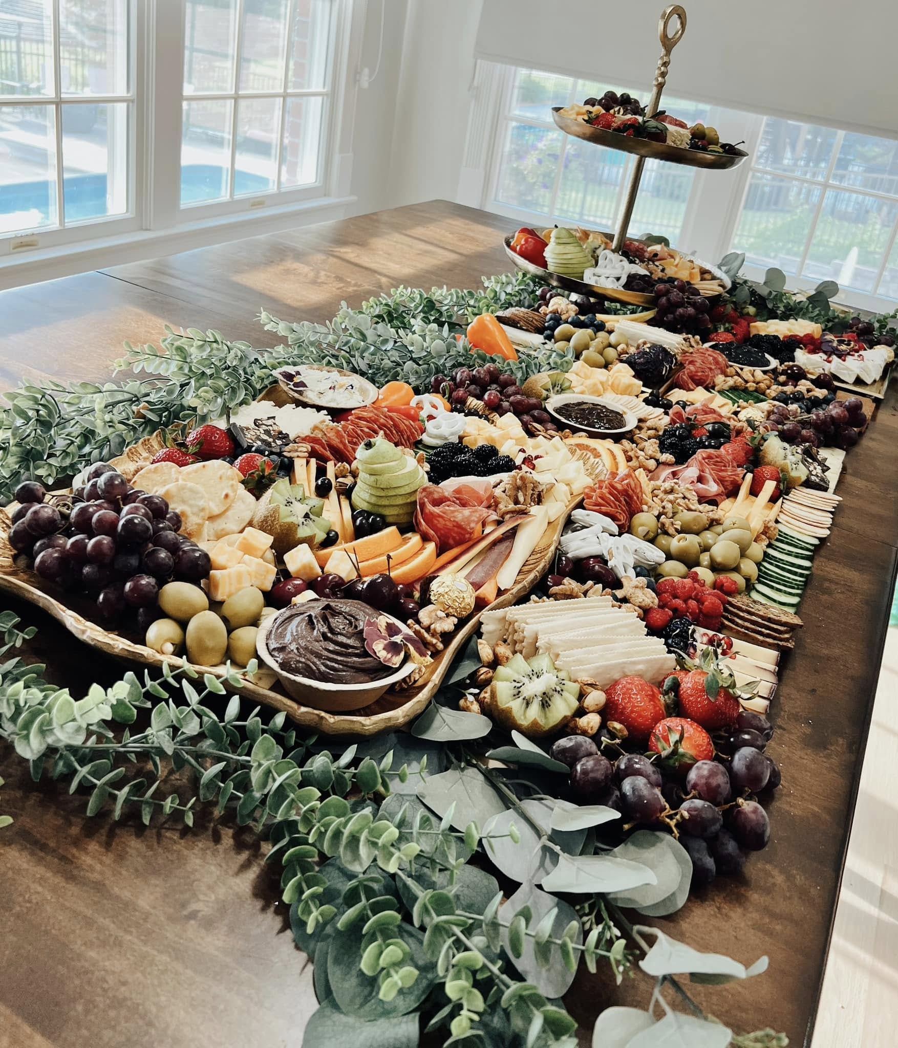 A long abundant grazing table with eucalyptus and gold trays in a sunlit dining room